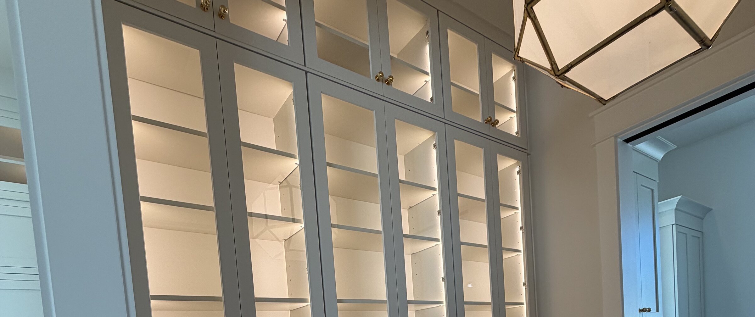 Butler's pantry with glass-front upper cabinets, integrated shelf lighting and brass knobs, built by Black Creek Cabinetry