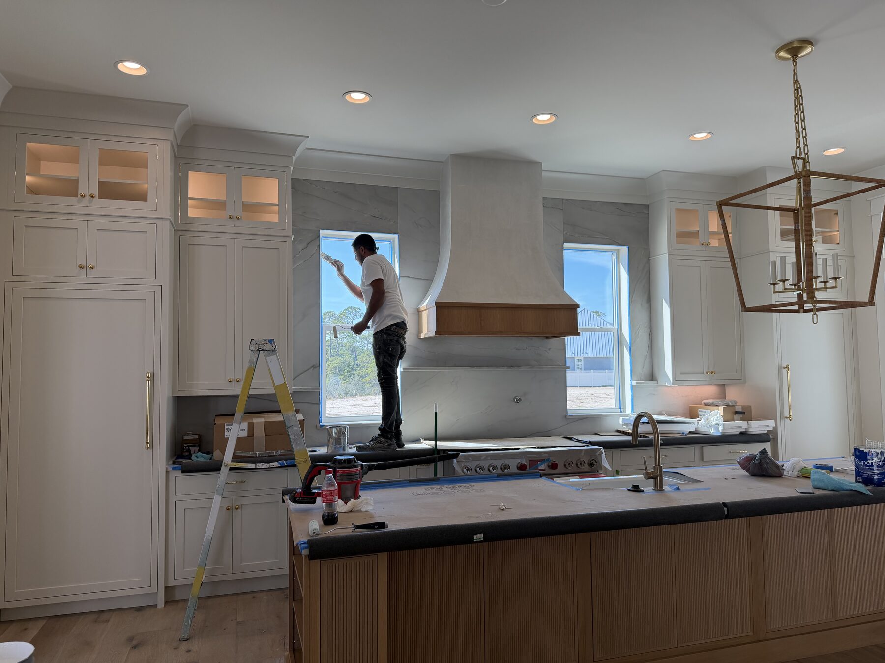 Large kitchen under installation with a custom range hood, marble slab backsplash, painted perimeter cabinets and a white oak island